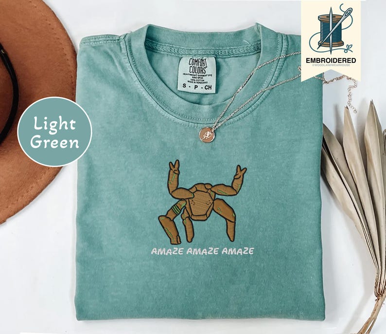 May include: Light green t-shirt with an embroidered brown crab design and the words "AMAZE AMAZE AMAZE" in white. The shirt has a crew neck and short sleeves. A silver necklace with a small pendant is visible.