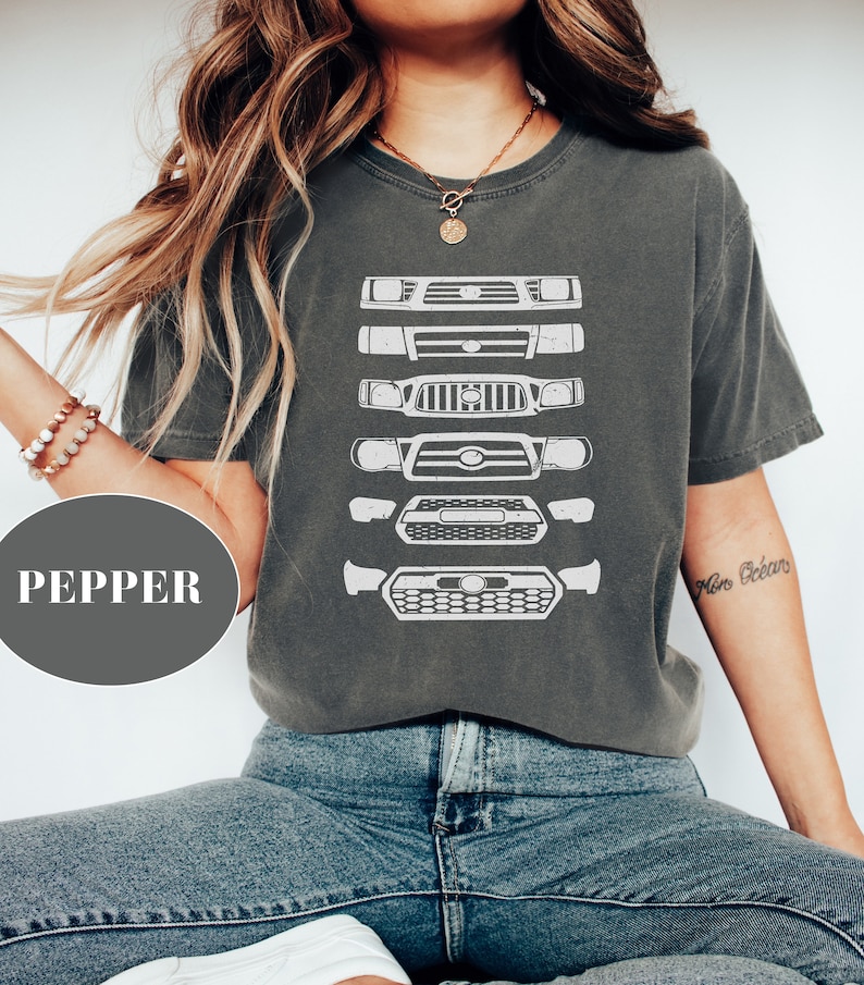 May include: A charcoal grey t-shirt with a graphic of a series of Toyota Tacoma front grilles printed in white. The shirt has a crew neck and short sleeves.