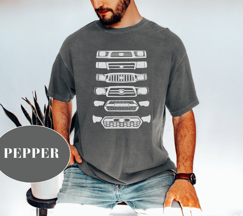 May include: A dark grey t-shirt with a white graphic of a series of Toyota Tacoma front ends. The graphic shows the evolution of the Tacoma front end from older models to newer models.
