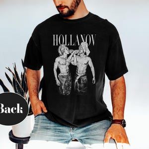 May include: Black t-shirt with the word "HOLLANOV" in white, above a black and white illustration of two figures. The figures are depicted in a vintage style, with one figure appearing to pour liquid into the other's mouth.