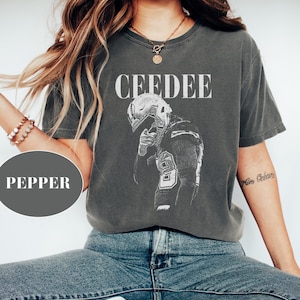 Könnte beinhalten: Dunkelgraues T-Shirt mit dem Wort "CEEDEE" über einer Schwarz-Weiß-Illustration eines Footballspielers. Das Shirt hat eine bequeme Passform und wird mit einer blauen Jeans kombiniert.