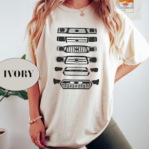 May include: A white t-shirt with a graphic of five different Toyota truck front grilles in black. The shirt is shown on a model wearing it with a pair of dark green biker shorts. The text "IVORY" is visible in the lower left corner of the image.