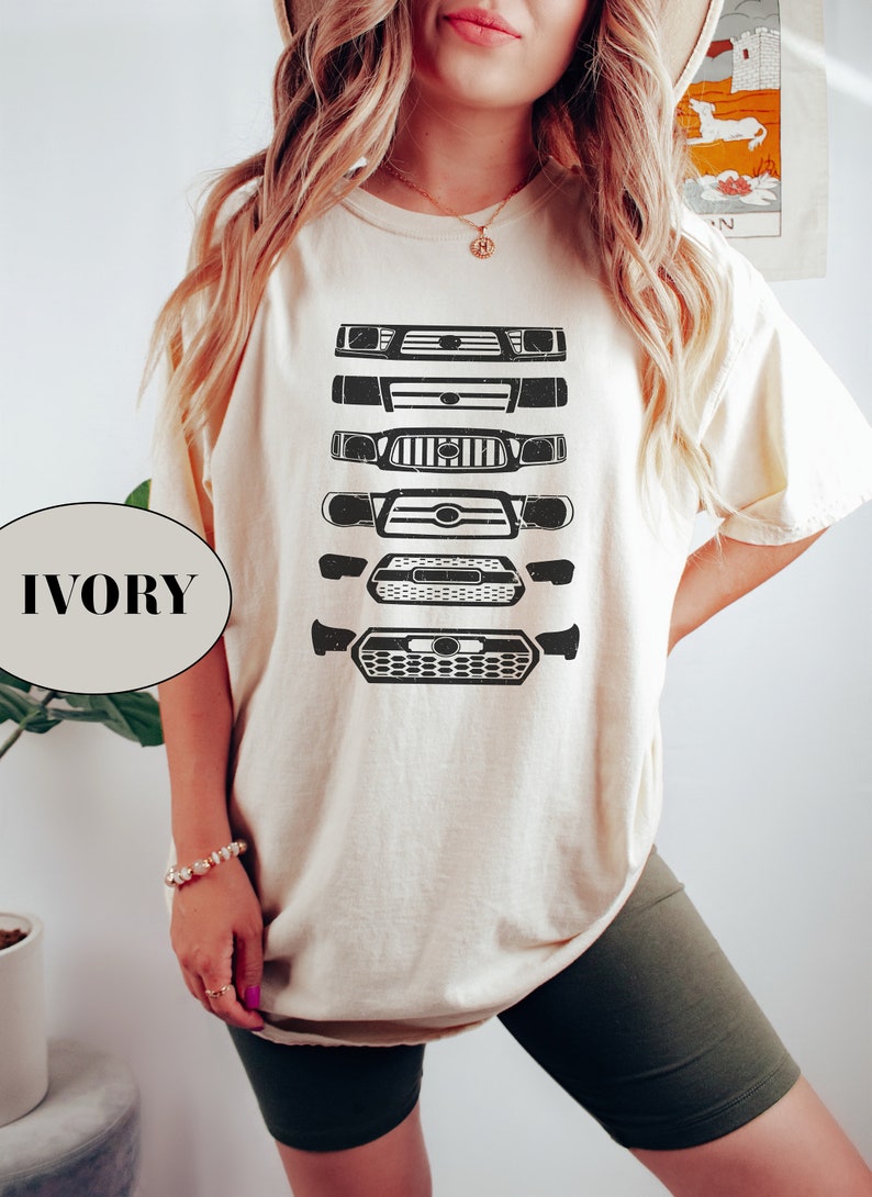May include: A white t-shirt with a graphic of five different Toyota truck front grilles in black. The shirt is shown on a model wearing it with a pair of dark green biker shorts. The text "IVORY" is visible in the lower left corner of the image.