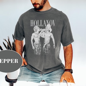 May include: Dark gray t-shirt with the word "HOLLANOV" above a black and white graphic of two figures. The figures are depicted in a vintage style. The shirt is worn by a person wearing blue jeans.