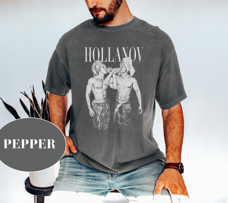 May include: Dark gray t-shirt with the word "HOLLANOV" above a black and white graphic of two figures. The figures are depicted in a vintage style. The shirt is worn by a person wearing blue jeans.
