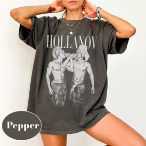 May include: Dark grey t-shirt with the word "HOLLANOV" in white block letters. The shirt features a graphic of two figures drinking from a bottle. The shirt is oversized and has short sleeves.