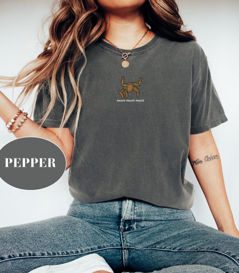 May include: A dark gray t-shirt with a small graphic of a stylized figure and the text "AMAZE AMAZE AMAZE" in brown. The shirt is paired with blue jeans. The word "PEPPER" is in a gray circle in the image.