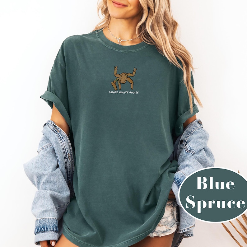 May include: Blue spruce-colored t-shirt with a cartoon crab graphic and the text "Amaze Amaze Amaze". The shirt is made of a soft, comfortable material.