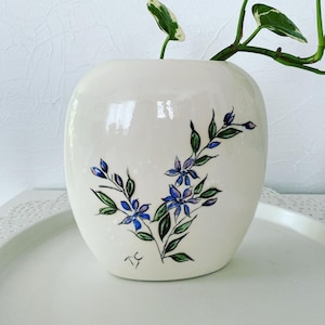 May include: White ceramic vase with a rounded shape, featuring a hand-painted design of blue and purple flowers with green leaves. The vase has a signature "TC" on the bottom.