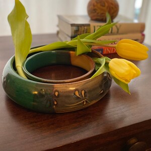 Peut inclure: Deux bracelets en céramique, l'un vert et l'autre bleu avec des accents blancs, sont empilés sur une table en bois. Un bouquet de tulipes jaunes repose sur les bracelets.