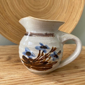 May include: A small, white ceramic jug with a brown handle and blue floral design. The jug has a brown stripe around the top and a brown and blue floral design on the side.