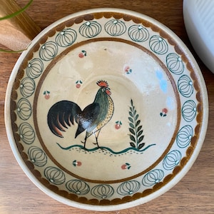 May include: A ceramic bowl featuring a rooster design. The bowl is white with brown and green accents. The rooster is black, green, and red. The bowl has a brown rim with a green and white pattern.