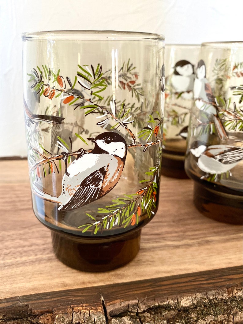 May include: Set of four vintage drinking glasses. Each glass features a detailed illustration of a bird perched on a branch with green needles and orange berries. The glasses have a brown base and a clear upper section.