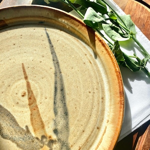 May include: A large, round ceramic plate with a brown rim and a beige centre. The plate has a hand-painted design of black and brown lines that resemble feathers.