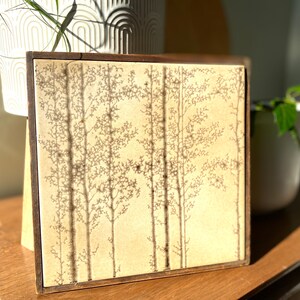 May include: A framed tile with a beige background and a brown wood frame. The tile features a design of stylised trees in a light brown colour.