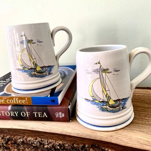 Peut inclure: Deux tasses en céramique blanche avec des motifs de voiliers bleus et jaunes. Les tasses sont posées sur deux livres, l'un intitulé "J'aime le café !" et l'autre intitulé "L'histoire du thé."