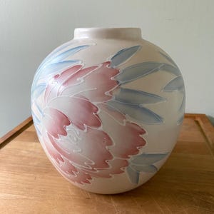 May include: A cream-coloured, round vase with a floral design. The design features a large pink and white flower with blue leaves. The vase is on a wooden surface.