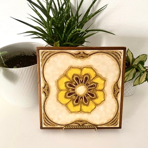 May include: A vintage ceramic tile featuring a yellow and brown floral design. The tile is framed in a wooden frame and has a gold stand.