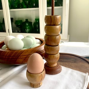 May include: A set of wooden egg cups, one with an egg inside, and a wooden egg holder with a tall wooden dowel in the centre. The egg cups and holder are made of light brown wood with a natural grain pattern.