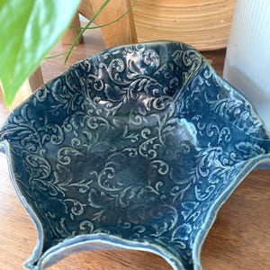 May include: A blue ceramic bowl with a decorative floral pattern. The bowl has a unique, irregular shape with a wide, shallow opening.