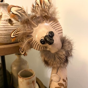 Peut inclure: Une figurine en céramique au design fantaisiste. La figurine a une barbe en fourrure brune, une coiffe en plumes brunes et blanches et des yeux noirs. La figurine est peinte avec des accents bruns, blancs et bleus.