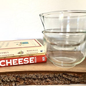 May include: Two clear glass mixing bowls stacked on top of each other on a wooden surface. The bowls are round and have a slight lip around the top. The wooden surface is a natural wood with a bark-like texture. Two books are stacked below the bowls. The top book is red and says "Cheese Book" in white letters. The bottom book is yellow and says "French Women Don't Get Fat Cookbook" in black letters.