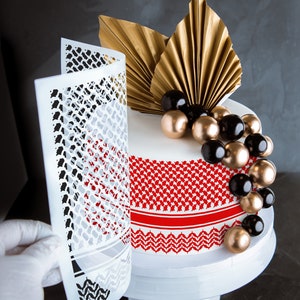 Puede incluir: Un pastel blanco decorado con un diseño estampado rojo y negro, acentos de pan de oro y bolas de chocolate negro y dorado. El pastel está sobre un soporte de pastel blanco.