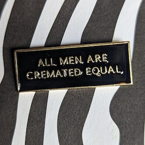 Könnte beinhalten: Schwarze Emaille-Anstecknadel mit goldenem Rand und dem goldenen Text "ALL MEN ARE CREMATED EQUAL". Die Nadel befindet sich vor einem schwarz-weiß gestreiften Zebramuster.