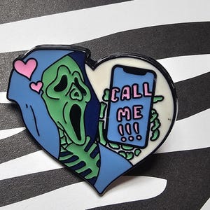 May include: A heart-shaped enamel pin featuring a green skeleton-like figure in a blue hood, with a screaming face. The figure holds a phone with the words "CALL ME" on the screen. Two pink hearts float above. The pin is set against a black and white zebra stripe background.