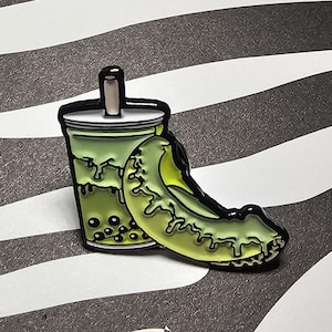 May include: A green and black enamel pin depicting a bubble tea cup and a pickle. The bubble tea cup has a white lid and a straw. The pickle is a light green colour with a dark green outline.
