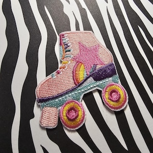 Op de afbeelding: Geborduurde patch van een roze rolschaats met een ster en regenboogaccenten. De patch heeft paarse, teal, gele en oranje details. De wielen zijn geel en roze. De achtergrond is een zwart-wit zebraprint.