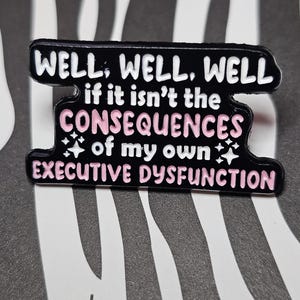 Peut inclure: Épinglette noire avec texte blanc et rose : « WELL, WELL, WELL if it isn't the CONSEQUENCES of my own EXECUTIVE DYSFUNCTION ». L'épingle a une forme unique, sur fond rayé noir et blanc.