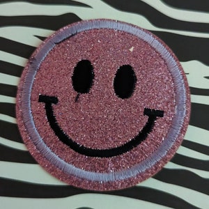 May include: Pink glitter smiley face patch with a black outline and a white border. The patch is on a black and white zebra print background.