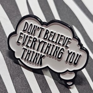 May include: A white enamel pin in the shape of a thought bubble with the words "DON'T BELIEVE EVERYTHING YOU THINK" in black lettering. The pin has a black outline and is set against a black and white striped background.