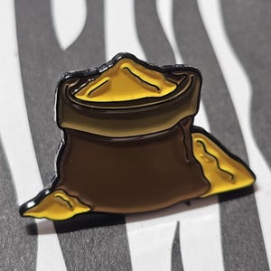 Könnte beinhalten: Ein brauner Emaille-Pin in Form eines Beutels, aus dem gelber Inhalt quillt. Der Pin hat einen schwarzen Umriss und ist vor einem schwarz-weiß gestreiften Hintergrund platziert. Der Beutel ist oben offen, wobei der gelbe Inhalt herausquillt.