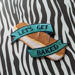 Può includere: Una toppa ricamata marrone e bianca con il testo "Let's Get Baked" in lettere blu turchese.