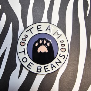 May include: A round, white enamel pin with the words "TEAM TOE BEANS" in black lettering. The center features a black cat paw with pink toe beans on a purple background. The pin is set against a black and white zebra print.