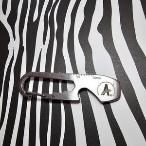 May include: A silver multi-tool featuring a carabiner clip, ruler markings, and a bottle opener. The tool has a wheat stalk logo and the letter "A". Measurements are in millimetres.