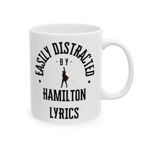 Puede incluir: Taza de cerámica blanca con el texto negro "EASILY DISTRACTED - BY - HAMILTON LYRICS". La taza presenta una silueta de una persona con el brazo levantado. La taza tiene un asa curva.