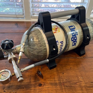 May include: A cylindrical Dräger Phoenix Firefighter breathing apparatus with a black handle and fittings. The tank is wrapped in a gray material with a white label. A pressure gauge and valve assembly are attached.