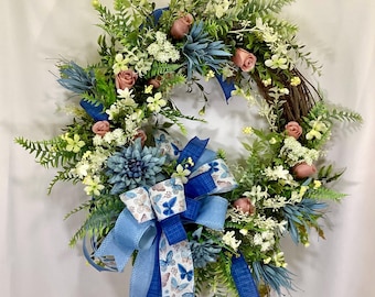 May include: A spring wreath with a blue and white butterfly ribbon bow. The wreath is made of grapevine and is decorated with pink roses, white flowers, and green leaves.