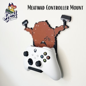 May include: A white Xbox controller mounted on a brown, cartoon-like wall mount shaped like a meatball. The mount has a black outline and two black hammer-shaped arms. The text "MEATWAD CONTROLLER MOUNT" is printed in black at the top of the image.