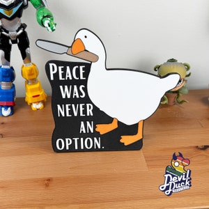 Op de afbeelding: Een witte gans met een mes in zijn snavel met de tekst "Peace was never an option." op een zwarte achtergrond. De gans staat op een zwarte ondergrond.