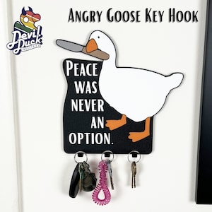 Puede incluir: Un colgador de llaves negro y blanco con forma de ganso enojado sosteniendo un cuchillo. El ganso es blanco con ojos negros y un pico negro. El texto en el colgador de llaves dice "Peace was never an option."