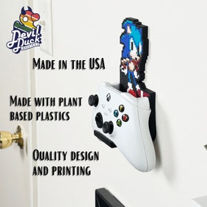 May include: A white Xbox controller is mounted on a wall with a 3D printed pixel art figure of Sonic the Hedgehog. The text "MADE IN THE USA" is visible above the controller. The text "MADE WITH PLANT BASED PLASTICS" is visible below the controller. The text "QUALITY DESIGN AND PRINTING" is visible below the Sonic figure.