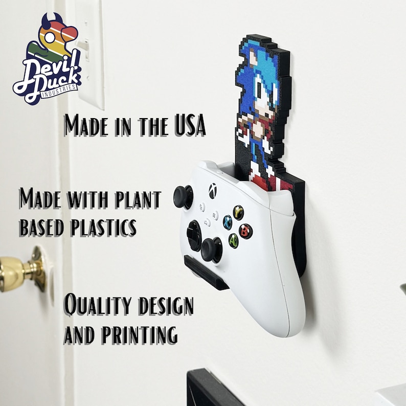 May include: A white Xbox controller is mounted on a wall with a 3D printed pixel art figure of Sonic the Hedgehog. The text "MADE IN THE USA" is visible above the controller. The text "MADE WITH PLANT BASED PLASTICS" is visible below the controller. The text "QUALITY DESIGN AND PRINTING" is visible below the Sonic figure.