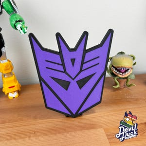 May include: Purple and black 3D printed Transformers logo. The logo is shaped like a robot face with a sharp, angular design.