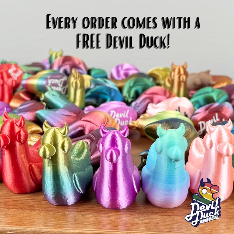 May include: A collection of colourful, iridescent, 3D printed duck figurines with horns. The ducks are arranged in a semi-circle on a wooden surface. The text "Every order comes with a FREE DEVIL DUCK!" is displayed above the ducks. The text "DEVIL DUCK INDUSTRIES" is displayed below the ducks.