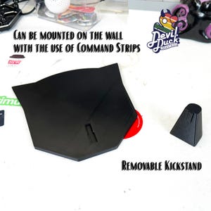 May include: Black plastic stand with a removable kickstand. The stand can be mounted on the wall using command strips. The text "Devil Duck Industries" is visible in the image.