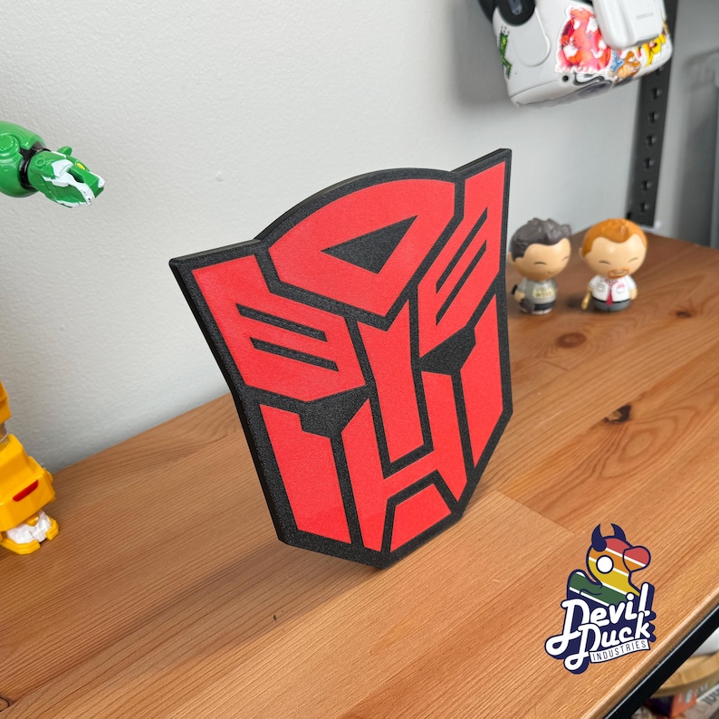 May include: Red and black Transformers Autobot symbol wall decor. The symbol is a stylized robot face with a red background and black outlines. The symbol is made of foam and is mounted on a wooden surface.